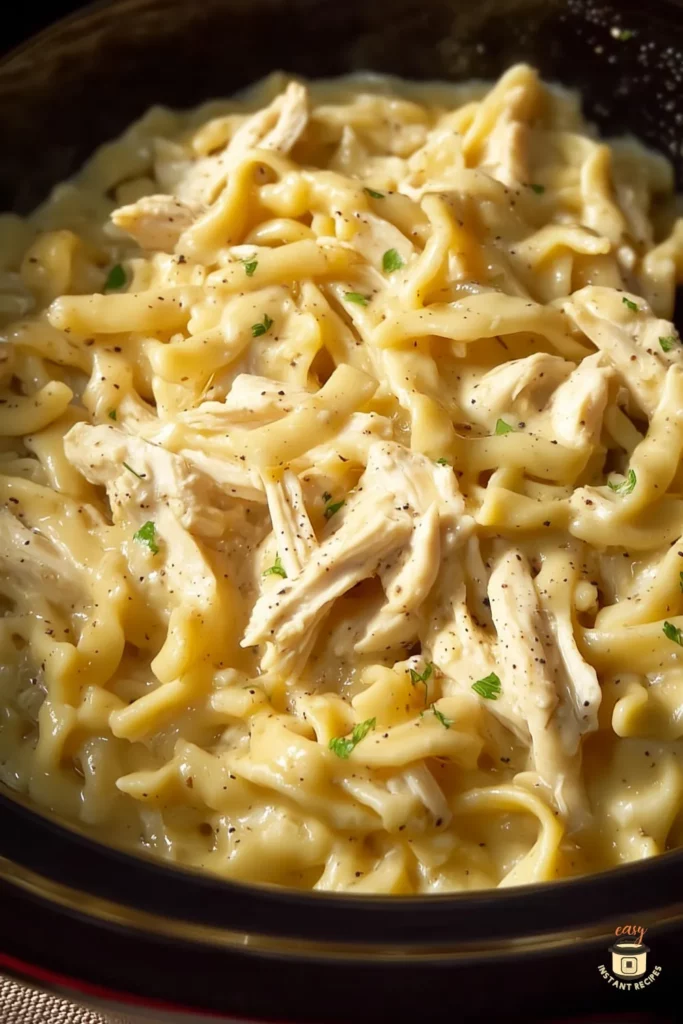 Close-up of creamy chicken and noodles cooked in the crockpot, showing tender shredded chicken mixed with thick egg noodles in a rich, seasoned sauce. Warm, comforting slow cooker dinner.